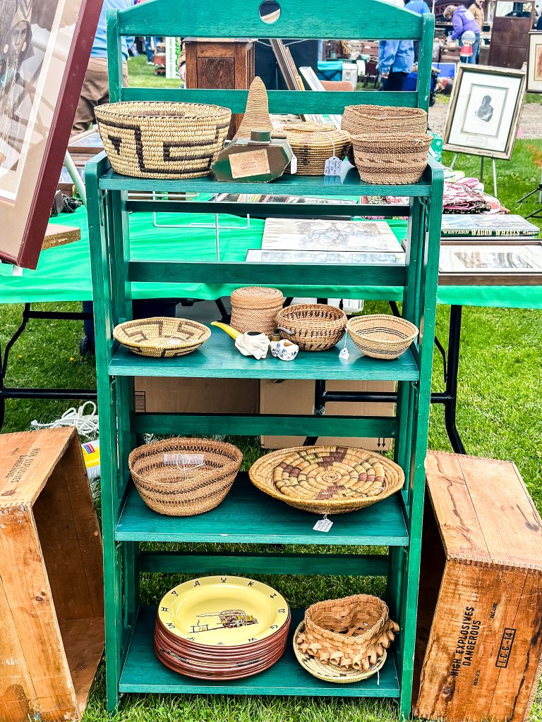 A green painted wooden rack displays a variety of woven baskets at a vintage market.
