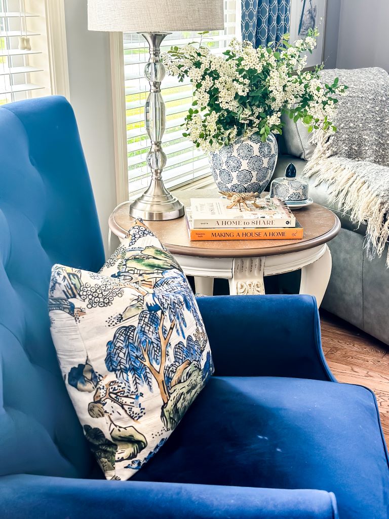 Blue velvet accent chair with a chinoiserie throw pillow next to a round white end table styled with stacked books, a silver lamp, and a blue and white ginger jar filled with white flowering branches.