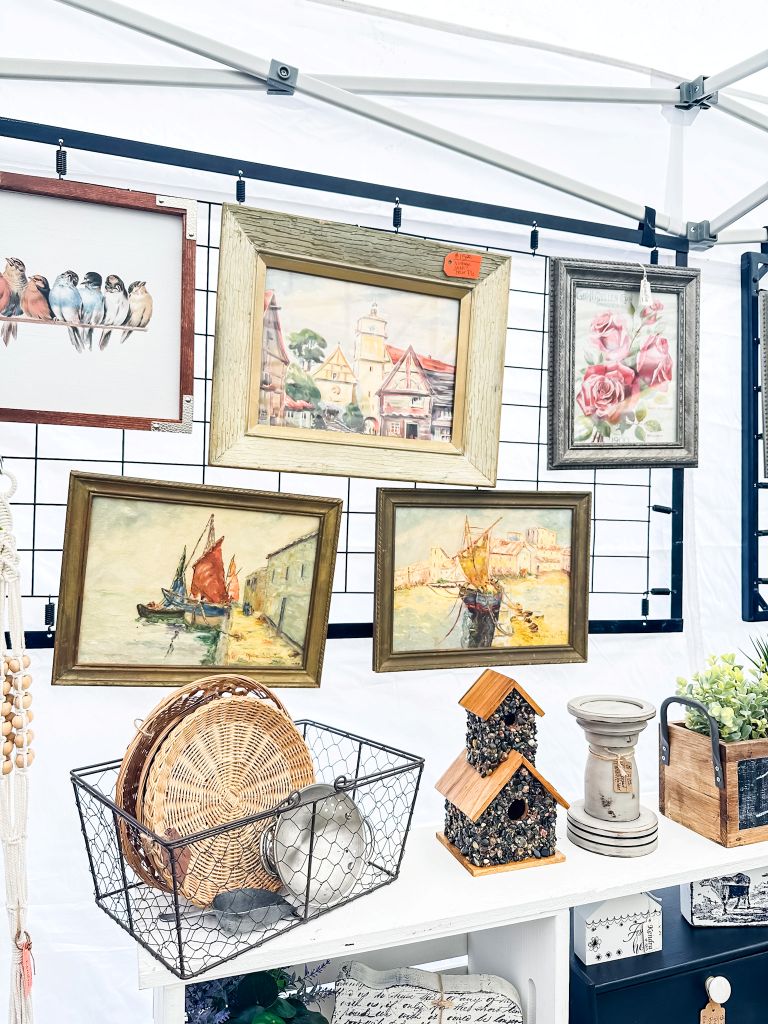 A display of framed vintage artwork, including landscapes, florals, and bird prints, at a vintage market.