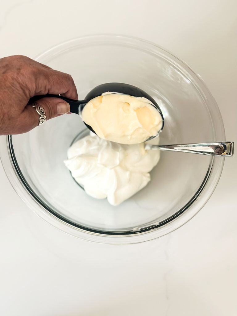 Adding a measuring cup of mayonnaise to a glass bowl of sour cream. The first step in making a creamy homemade dill dip recipe with fresh garden herbs.