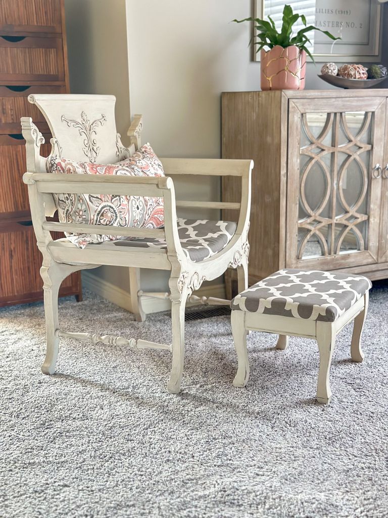 Ornate light-colored wooden accent chair with a patterned cushion and matching ottoman on a grey carpet. The furniture has a distressed or antique finish, complementing the classic decor.