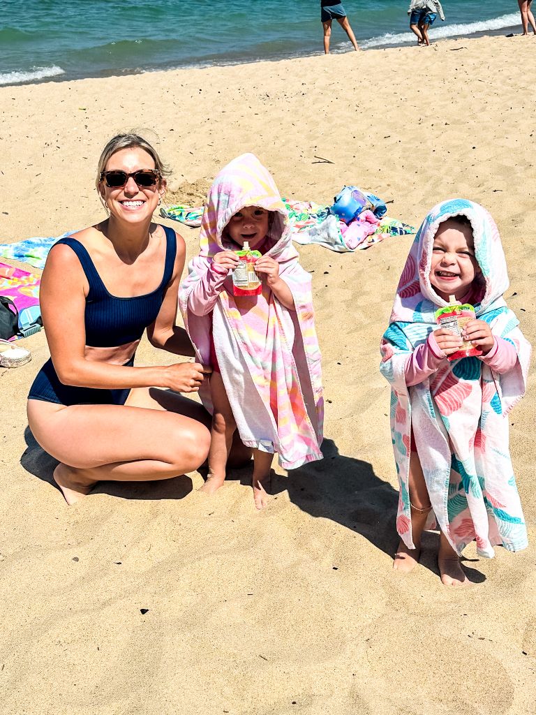 Mom posing with twin toddler girls in hooded beach towels enjoying juice pouches on a Lake Michigan beach.