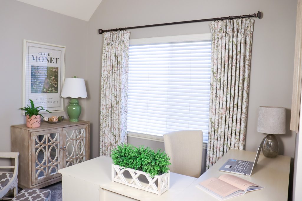 Bright and cozy home office with floral pleated curtains, from TheHue, a white desk with greenery, a light wood cabinet with mirrored doors, and soft neutral decor including a green lamp and blush pink planter.