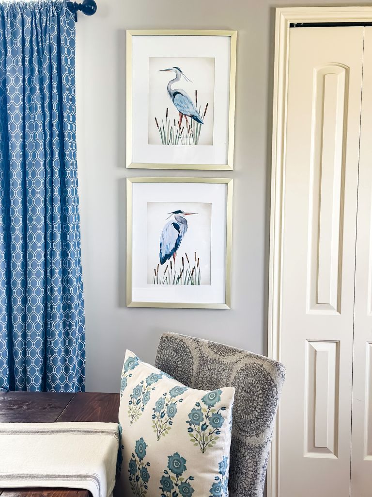 Framed blue heron watercolor art prints in gold frames hung vertically in a dining room with blue geometric curtains, a floral cottage pillow, and dark wood dining table.