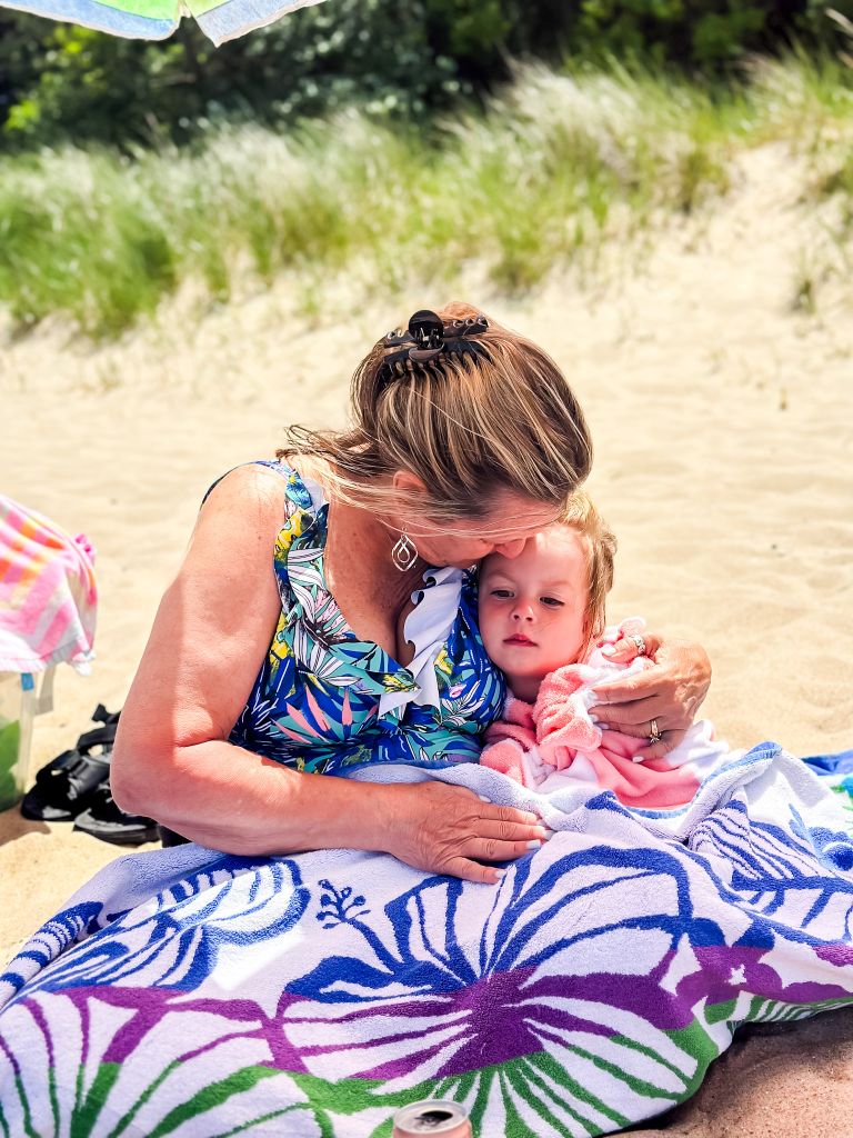 Grandmother cuddling a sleepy toddler wrapped in a pink towel on a Lake Michigan sandy beach.