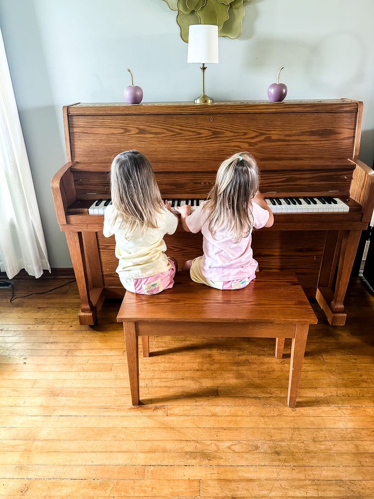 Two little girls sitting together on a piano bench playing an upright piano.