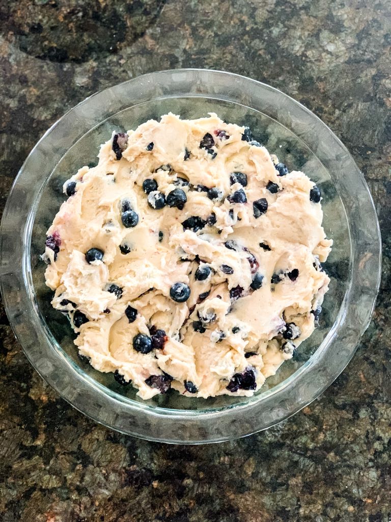 Overhead view of a thick, light-colored batter with blueberries mixed in, spread evenly in a clear glass pie dish.