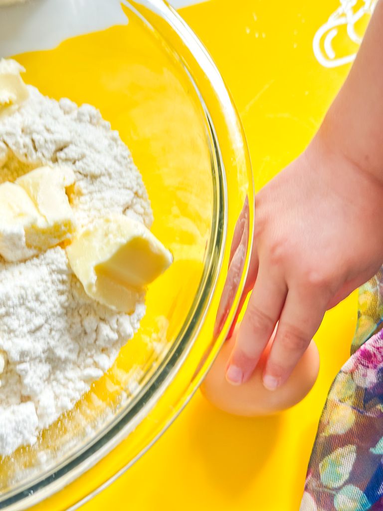 A child's hand holding an egg next to a glass bowl containing cookie mix and pieces of butter, all on a bright yellow cutting mat.