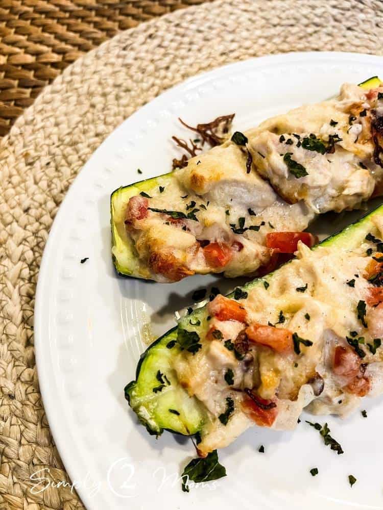 Two baked zucchini boats filled with chicken alfredo, tomatoes, and cheese, garnished with fresh herbs.