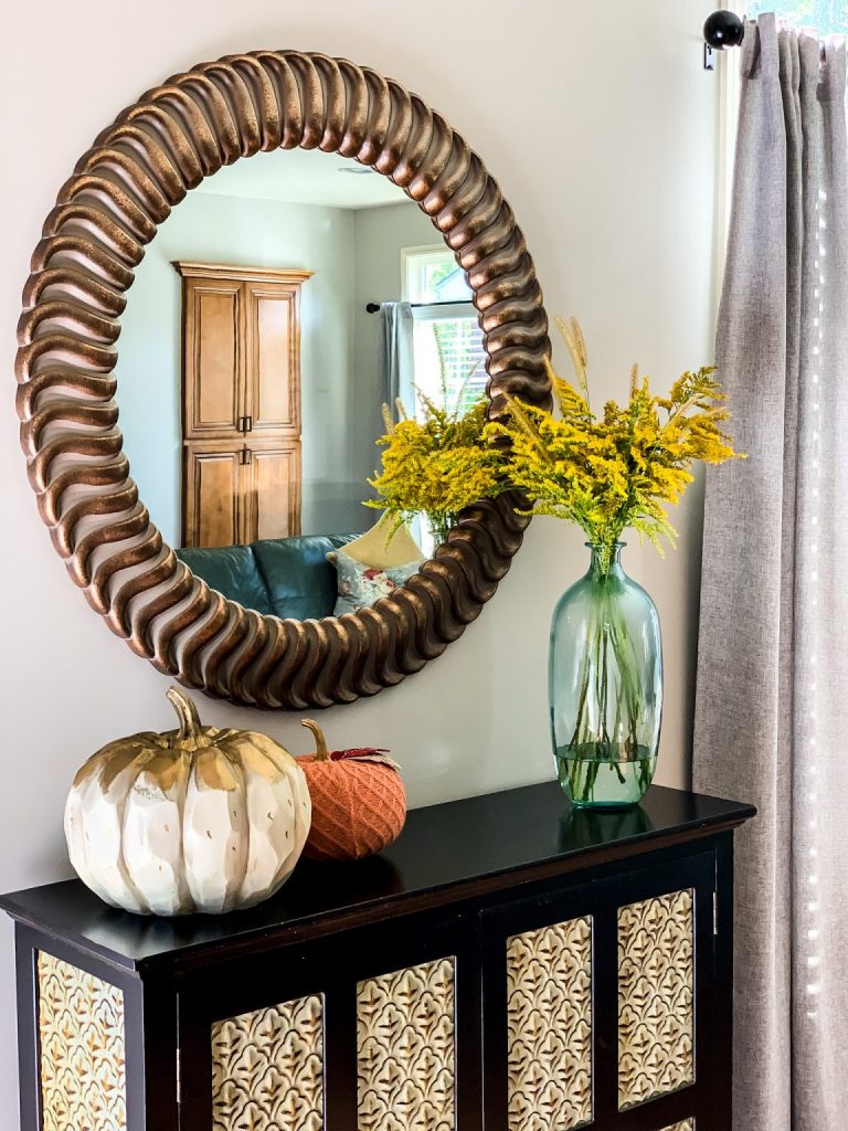 An autumn console table vignette featuring a large, round mirror, painted pumpkins, and a light blue vase filled with a bouquet of foraged goldenrod.