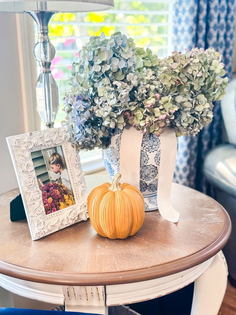 A fall side table vignette featuring a blue and white ceramic vase with a large bouquet of dried hydrangeas, a decorative pumpkin, and a white ornate picture frame.