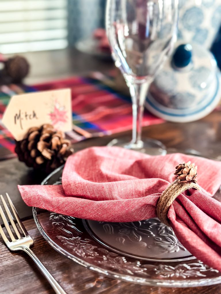 A rustic fall table setting featuring a DIY jute twine and pinecone napkin ring on a red cloth napkin, with a pinecone place card and name tag.