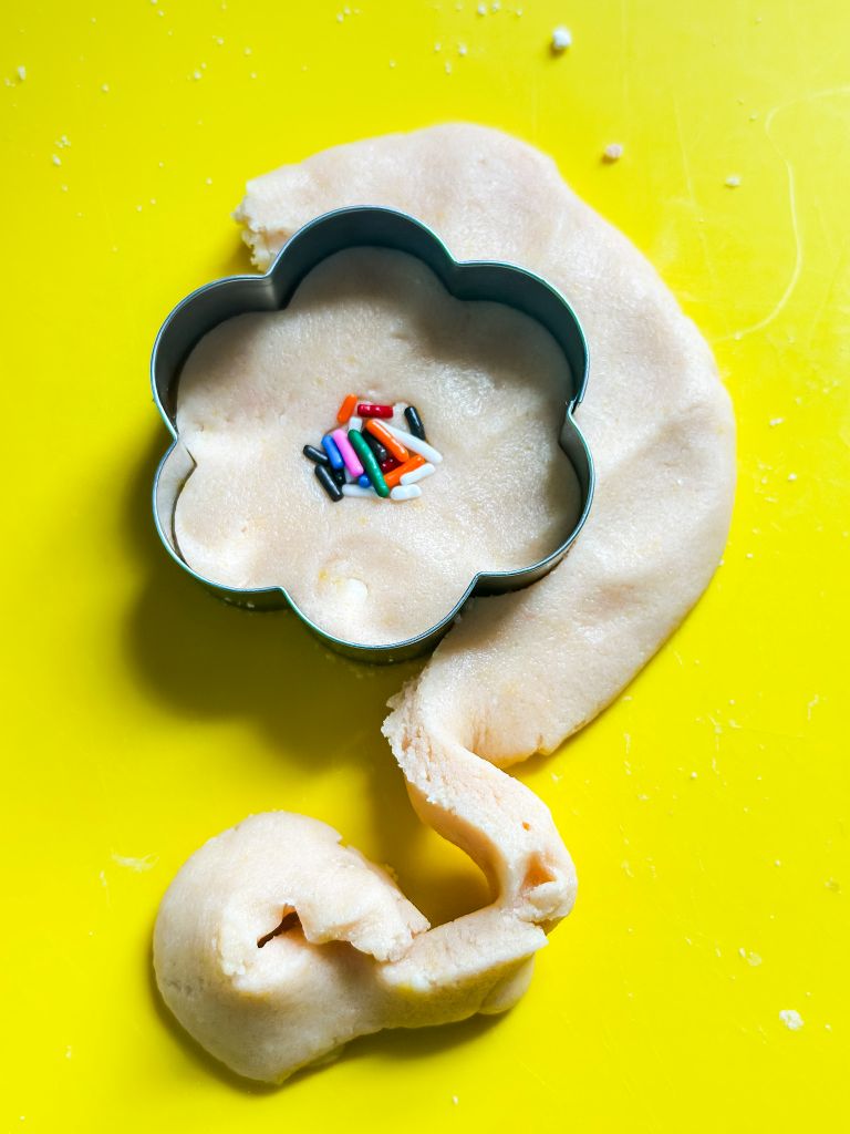Flower-shaped cookie cutter pressing into sugar cookie dough with colorful sprinkles in the center, on a bright yellow cutting mat.