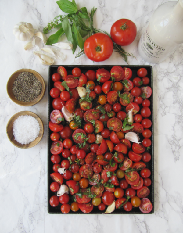 An overhead view of a sheet pan filled with cherry and plum tomatoes, garlic cloves, and fresh basil, ready for roasting. Salt, pepper, and a bottle of olive oil are also on the counter.