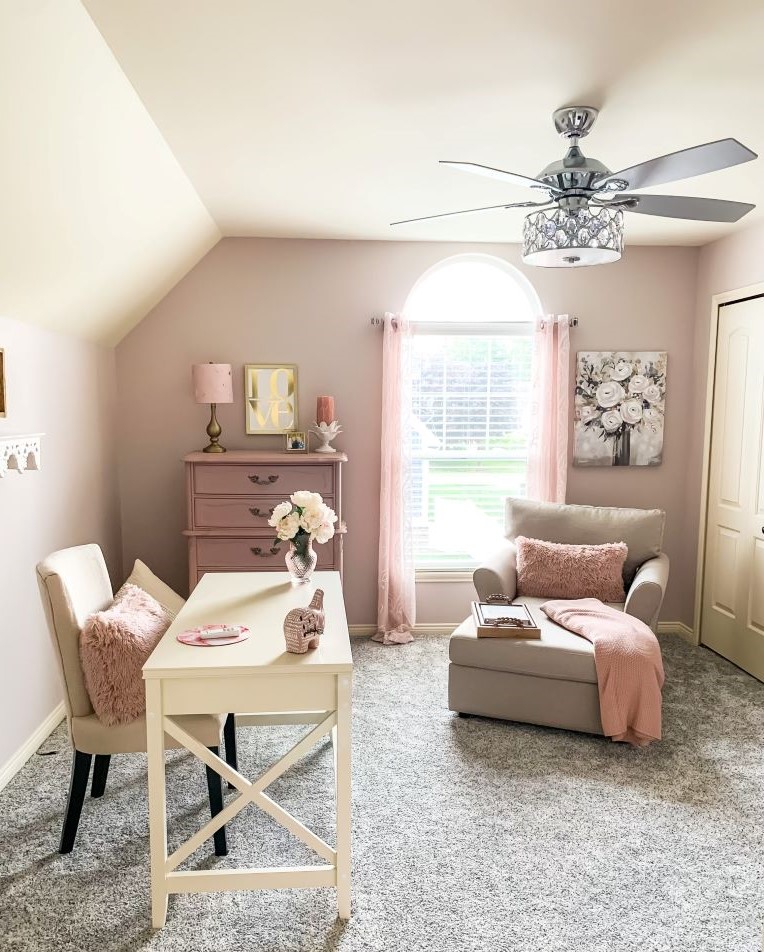 A bright and airy home office or she-room with a white desk and a pink dresser against a light pink wall. A plush cream colored chaise lounge sits by a large window with sheer pink curtains.