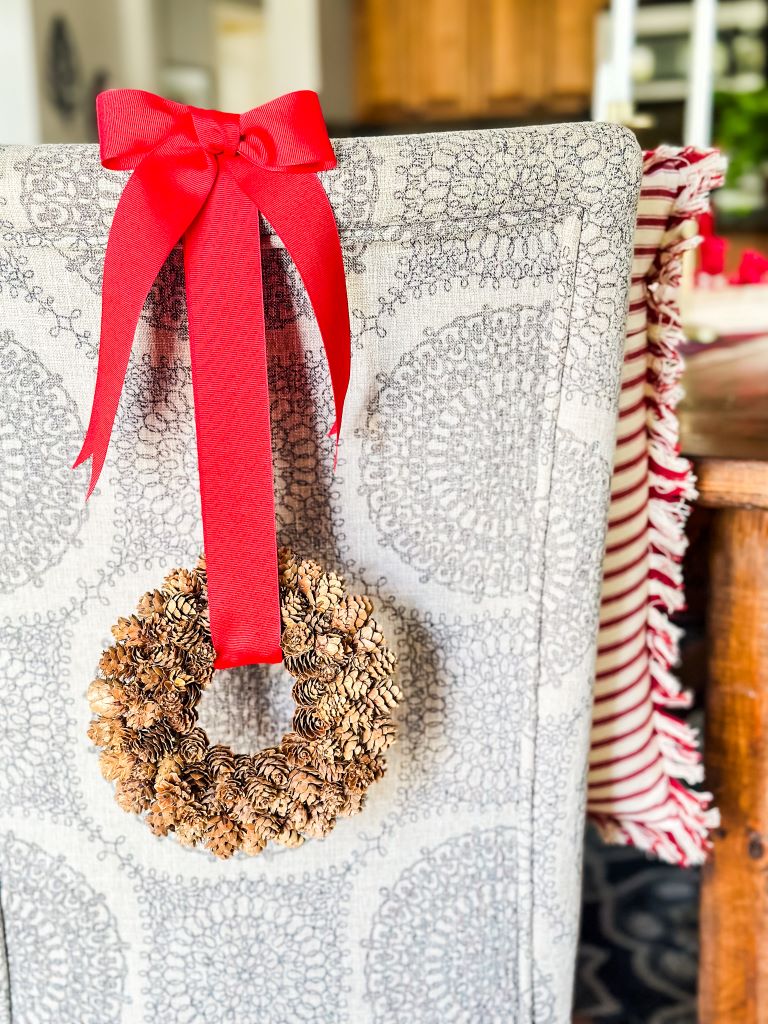 A small, handmade wreath made from tiny pinecones, hanging from a red ribbon on the back of a dining chair.