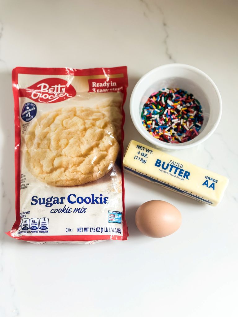 Betty Crocker Sugar Cookie mix, butter, a white bowl with sprinkles and an egg sitting on a white quartz counter. 
