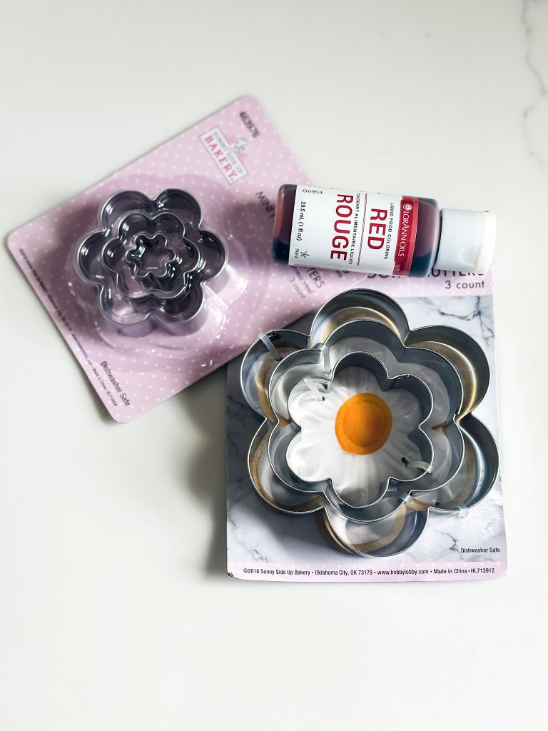 Two packages of cookie cutters shaped like flowers in different sizes and a bottle of red food coloring. 