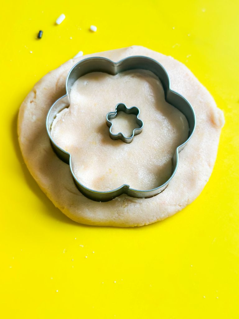 Two fluted metal flower cookie cutter firmly into a pale, raw sugar cookie dough that has been rolled out on a bright yellow silicone baking mat or cutting board. 