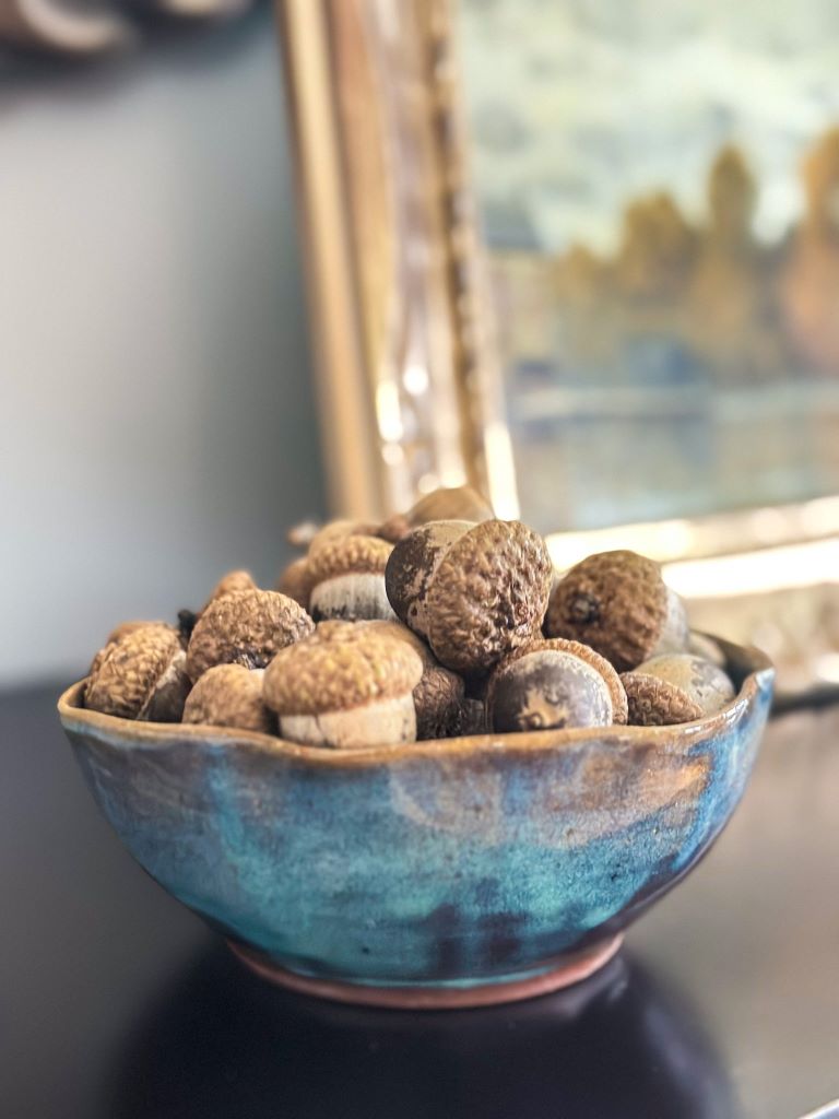 A blue ceramic bowl on a counter with acorns in it. A fall picture in a gold frame is behind it. 