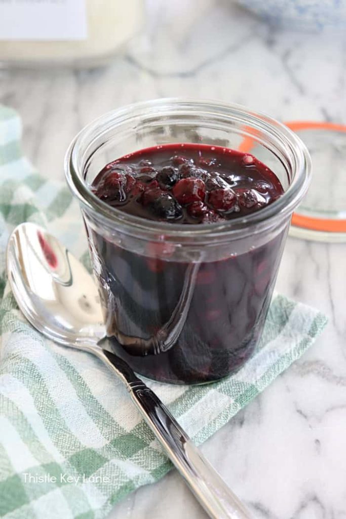 A close-up of a glass jar filled with homemade blueberry compote, with a silver spoon resting next to it on a checkered napkin.