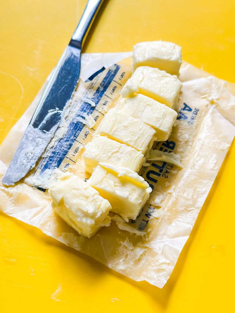 A stick of butter, partially unwrapped, that has been cut into cubes on a yellow cutting board, with a knife resting on the butter wrapper.