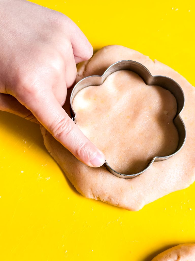 A person's hand pressing a metal flower cookie cutter into a light-colored sheet of unbaked cookie dough on a bright yellow surface.
