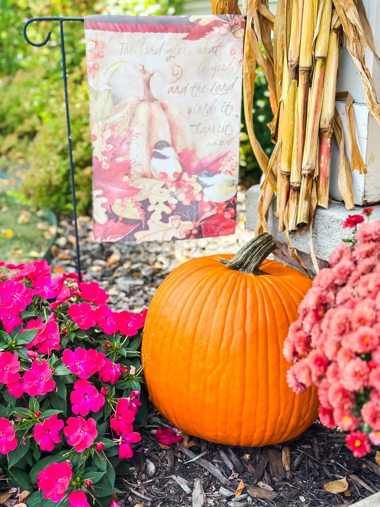 Fall garden flag with a Psalm 85:12 Bible verse, pumpkins, and chickadees, displayed near cornstalks and pink impatiens on the porch.