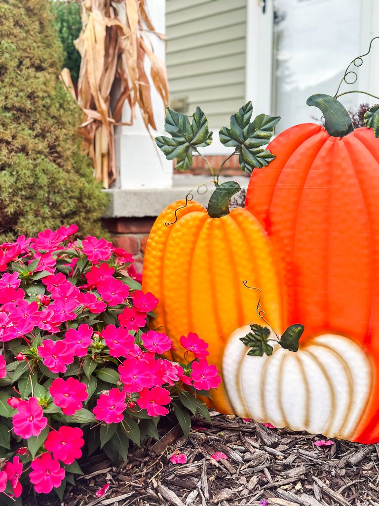 Metal pumpkin decor in orange and white nestled among fuchsia New Guinea impatiens on a fall porch, with cornstalks in the background.