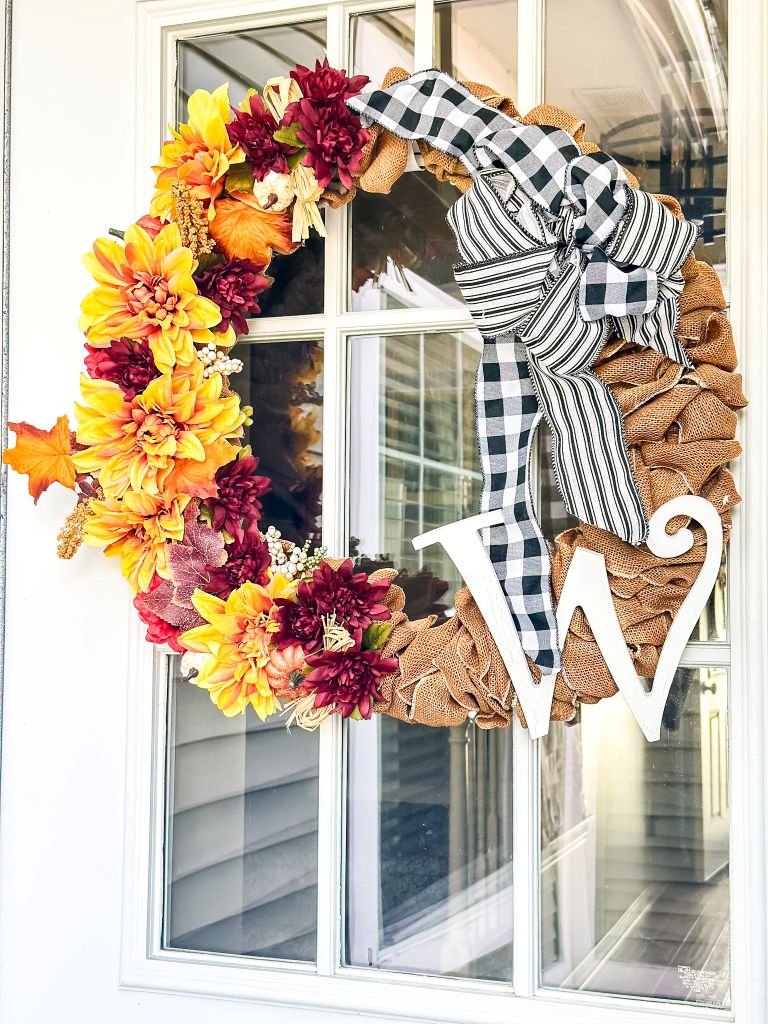 Rustic burlap fall wreath with black and white buffalo check ribbon, yellow and burgundy flowers, and a large white monogram on a porch door.