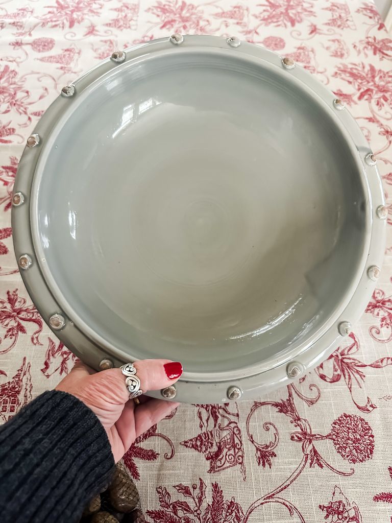 Overhead close-up of a hand with red nail polish holding a large, shallow ceramic vessel in a soft, dusty blue-gray color. The bowl features a detailed rim with raised, button-like decorative studs. The vessel rests on a patterned red and white toile tablecloth.