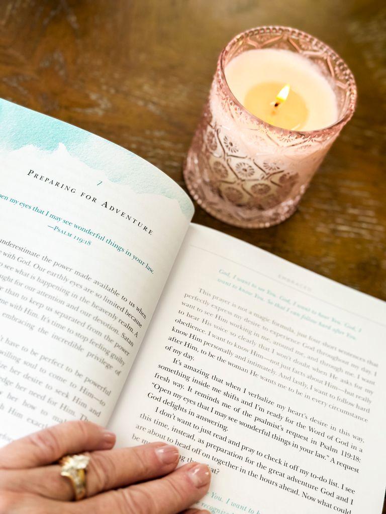 A cozy morning ritual in a traditional cottage home featuring a lit candle in a pink hobnail glass jar sitting next to an open devotional book titled Embraced by Lysa TerKeurst on a rustic wood table.