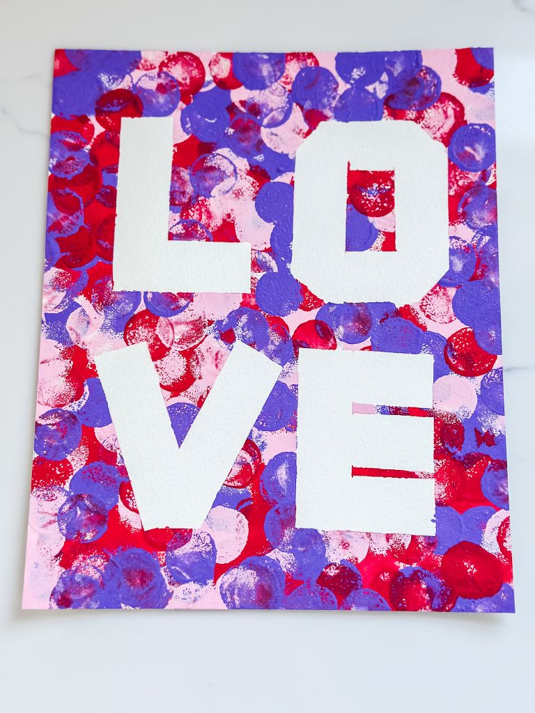 Completed Valentine’s Day tape-resist dot art spelling 'LOVE' on canvas paper, featuring vibrant pink, red, and purple paint dabs.