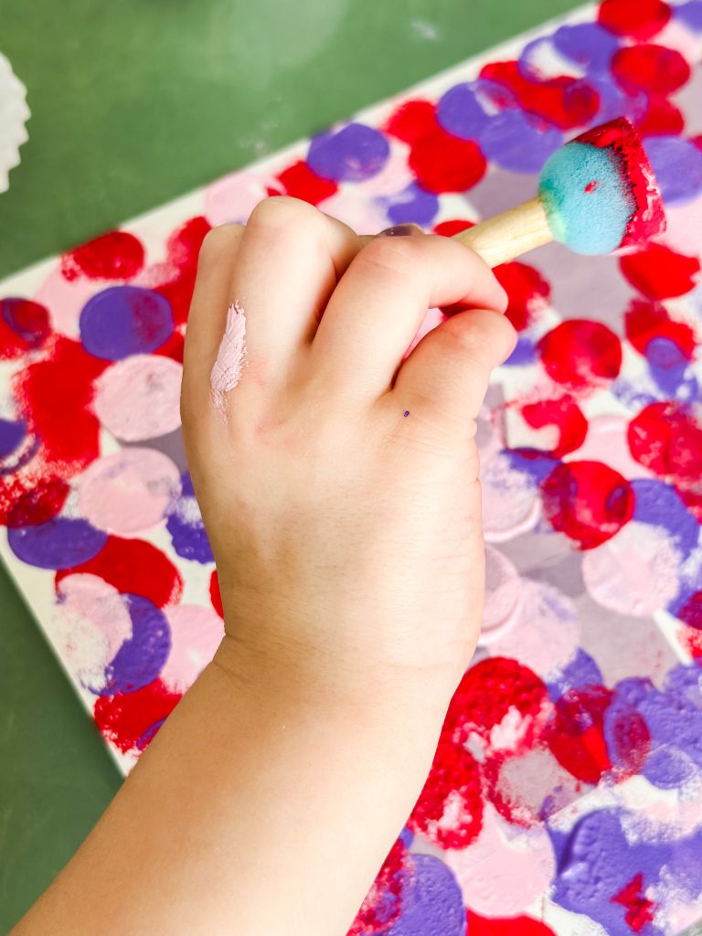 A toddler's hand using a round sponge foam brush to dap red and pink paint onto a canvas for a Valentine's Day process art project.