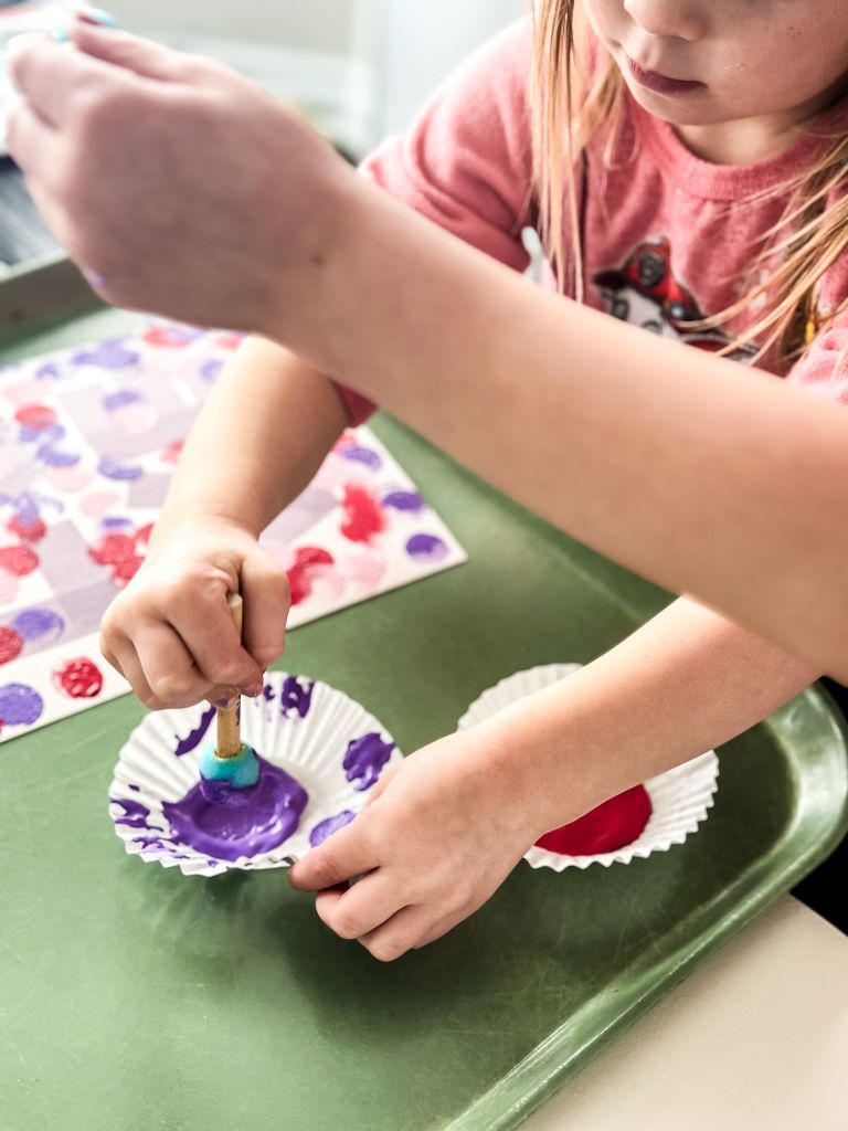 A child using a sponge dauber to dip into purple paint held in a cupcake liner, working on a Valentine’s Day tape-resist canvas art project.