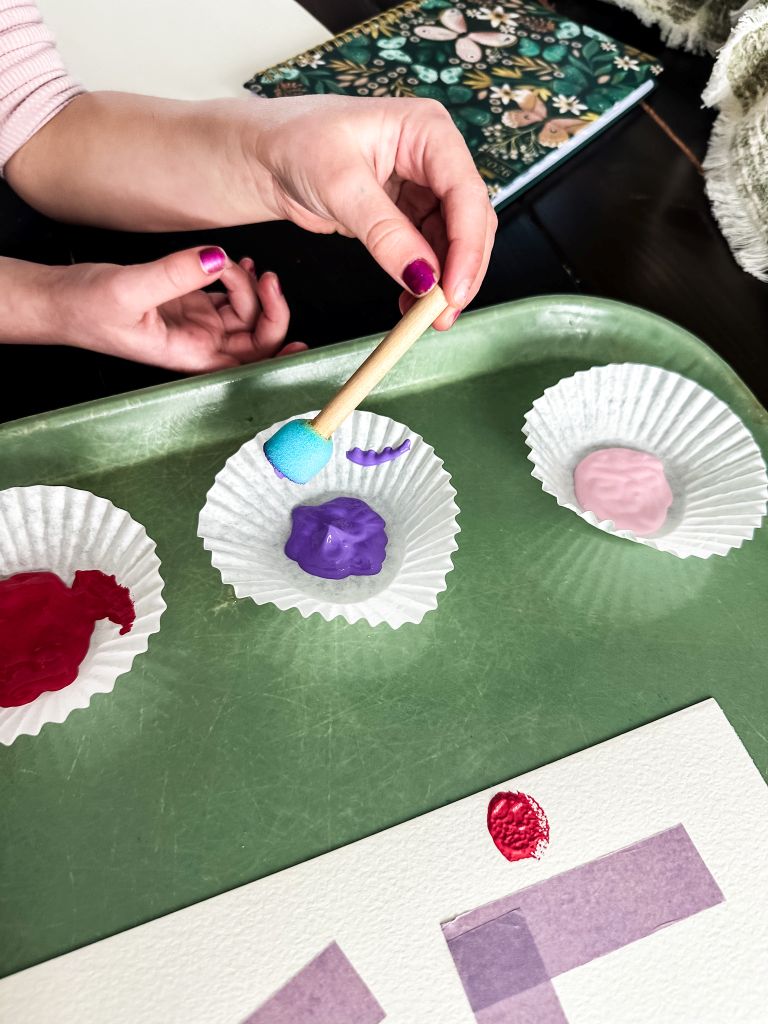 Pink, red, and purple paint in white cupcake liners on a green tray, being used with a sponge dauber for a Valentine's Day kids' craft.