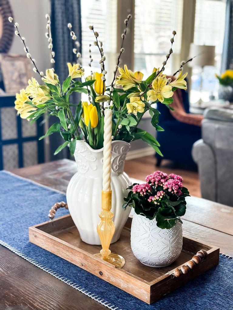 Spring cottage dining table decor featuring a white ironstone pitcher filled with yellow tulips and pussy willow branches, a small pink flowering plant, and a yellow glass taper candle holder on a wooden beaded tray.