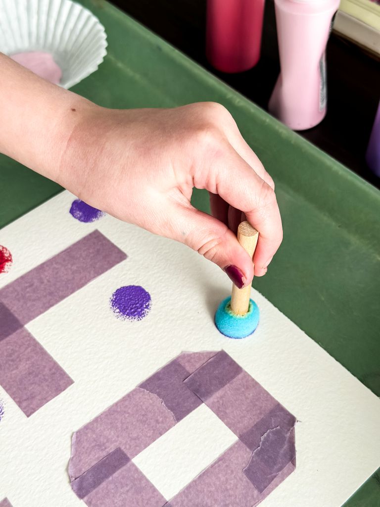 A hand using a blue sponge dauber to apply a purple paint dot next to masking tape letters on a white canvas for a Valentine's Day craft.