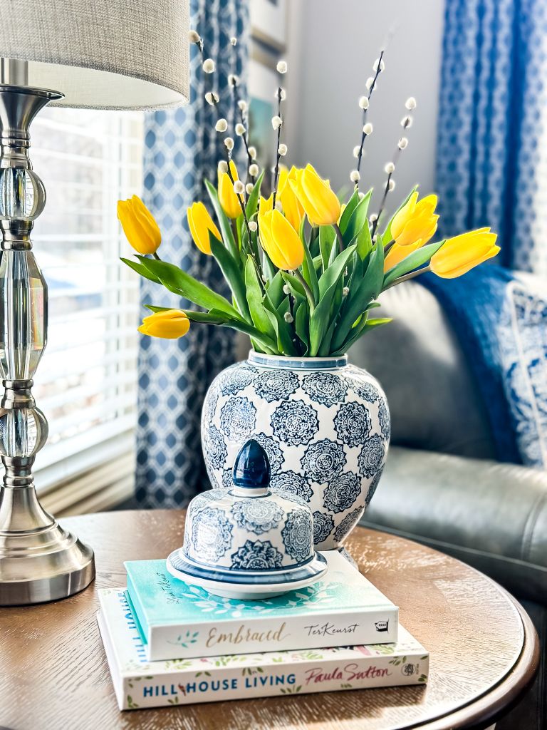 Cottage living room side table featuring a blue and white patterned ginger jar filled with yellow tulips and pussy willow stems, styled on a stack of books including 'Embraced' by Lysa TerKeurst and 'Hill House Living' by Paula Sutton.