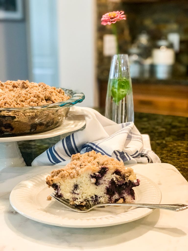 A slice of fresh blueberry buckle coffee cake on a white plate with a fork, styled in a cottage kitchen with a blue striped linen napkin, a white cake stand, and a single pink flower in a glass vase.