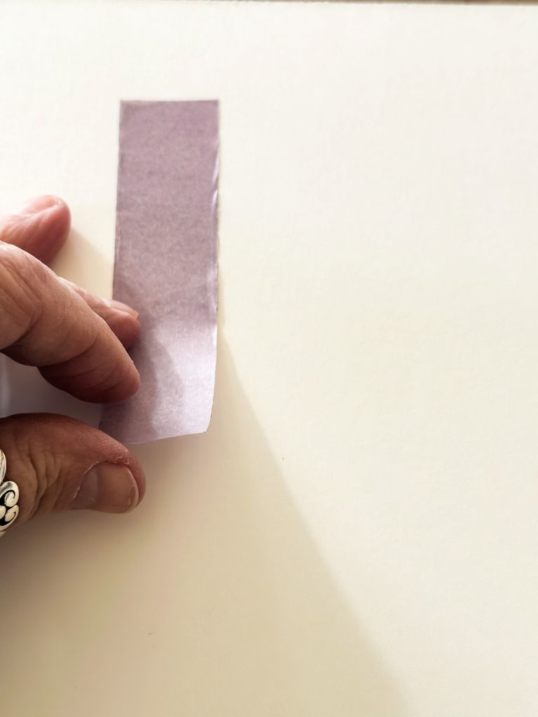 A hand carefully placing a strip of purple masking tape onto white canvas paper to create a letter for Valentine’s Day tape-resist art.