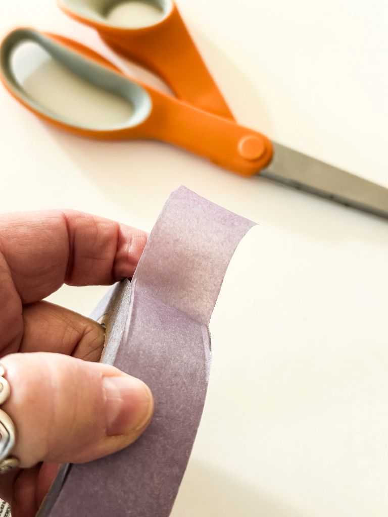 Close-up of a hand holding purple painter's tape with orange scissors in the background, preparing a tape-resist Valentine's Day craft.