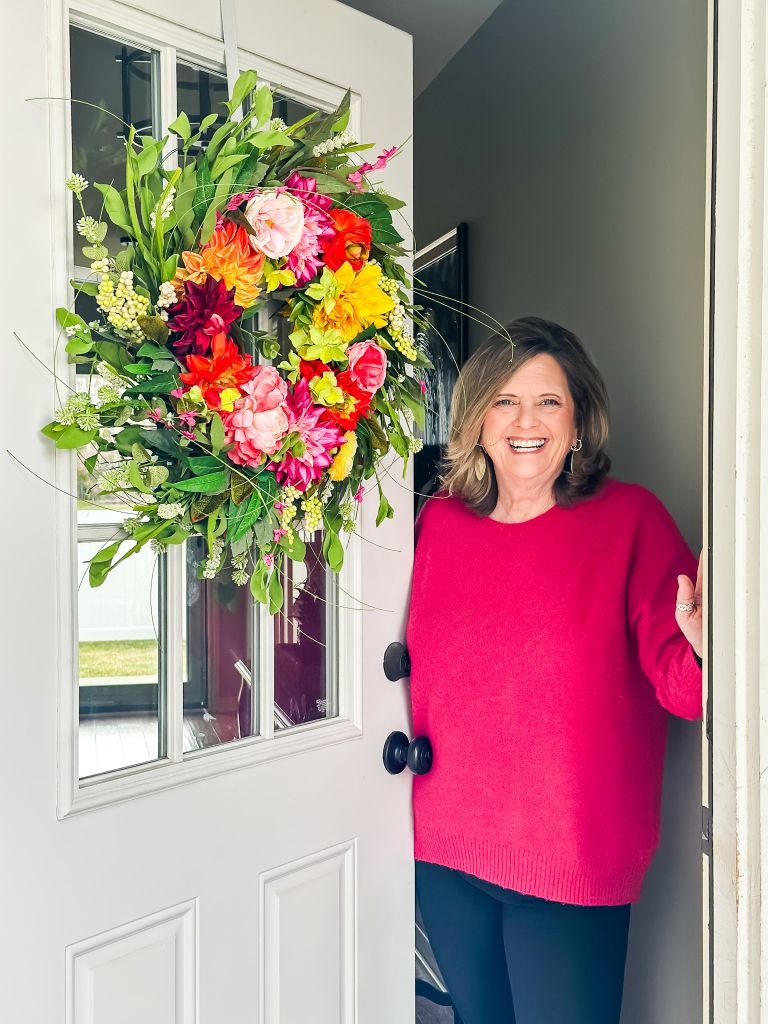 A smiling Kim Waldorf standing in the open doorway of her cottage, wearing a bright pink sweater next to a large, vibrant spring floral wreath with pink, orange, and yellow dahlia blossoms and lush greenery.