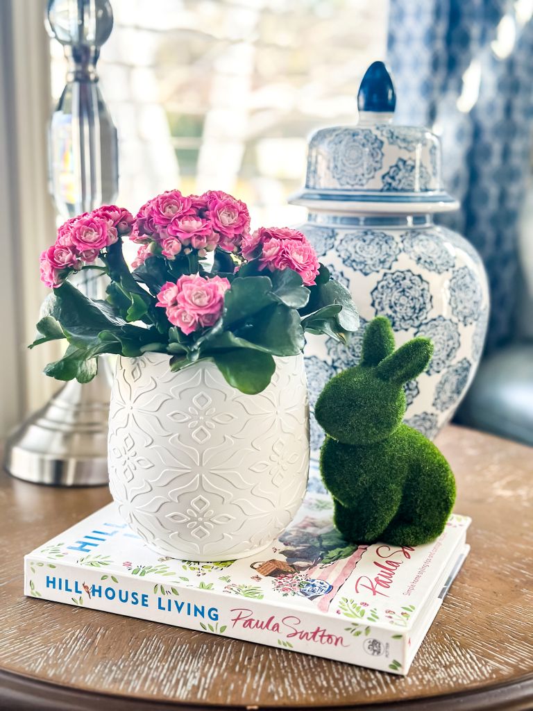 A spring cottage living room vignette featuring vibrant pink kalanchoe flowers in a white textured pot, resting on a "Hill House Living" coffee table book alongside a green moss bunny and a blue and white ginger jar