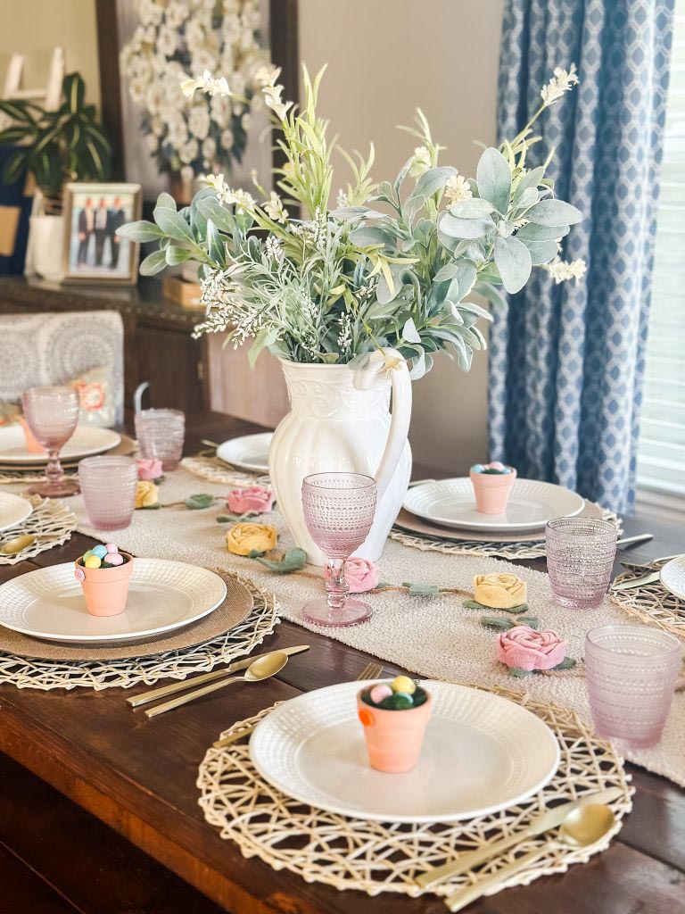 A complete view of a spring tablescape featuring a white ceramic pitcher centerpiece with lamb's ear and wildflowers, set on a dark wood table with pink hobnail glassware, gold flatware, and individual mini terra cotta candy pots at each place setting.