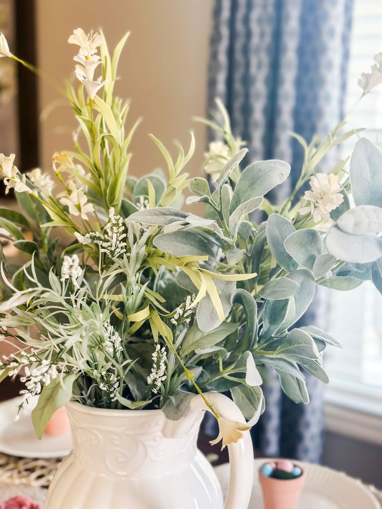 A beautiful spring table centerpiece featuring a white ceramic pitcher filled with a lush arrangement of lamb's ear greenery, delicate white wildflowers, and soft green stems, set against a background of a classic cottage dining room.