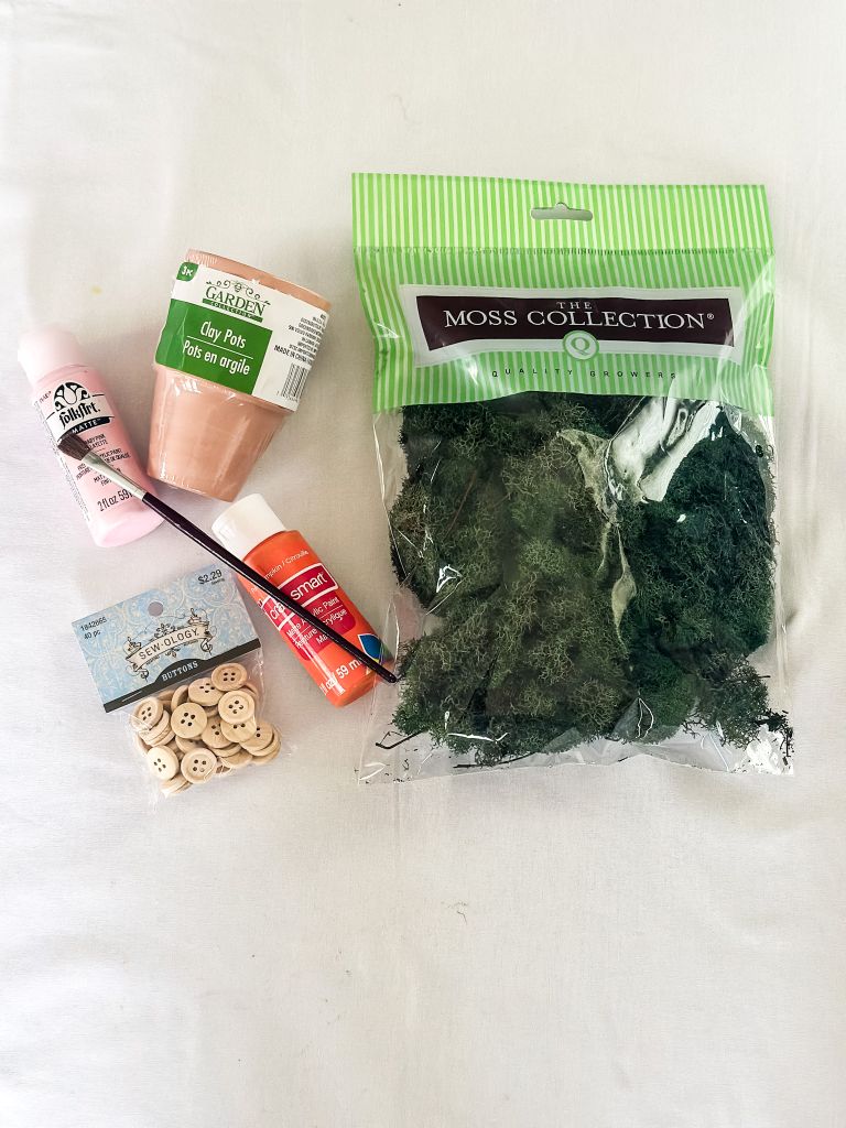 A flat lay of DIY craft supplies for a spring project, including small terra cotta clay pots, a bag of preserved green moss, pink and orange acrylic craft paint, a paintbrush, and a small package of natural wooden buttons on a white background.