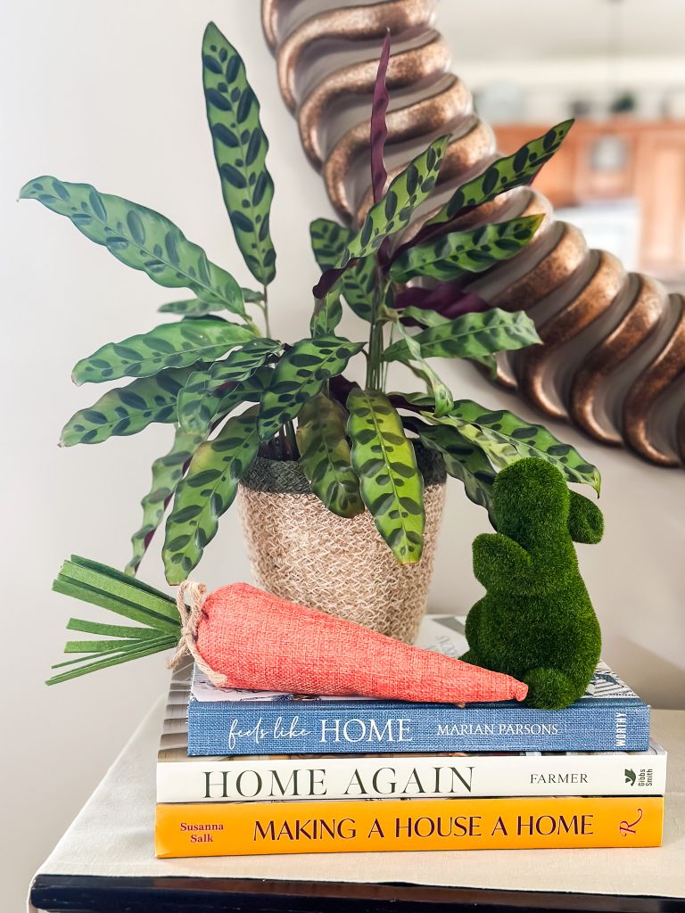 A traditional console table display featuring a stack of home decor books including "Feels Like Home" and "Home Again," topped with a large fabric burlap carrot and a green moss bunny next to a patterned Calathea plant in a woven basket.