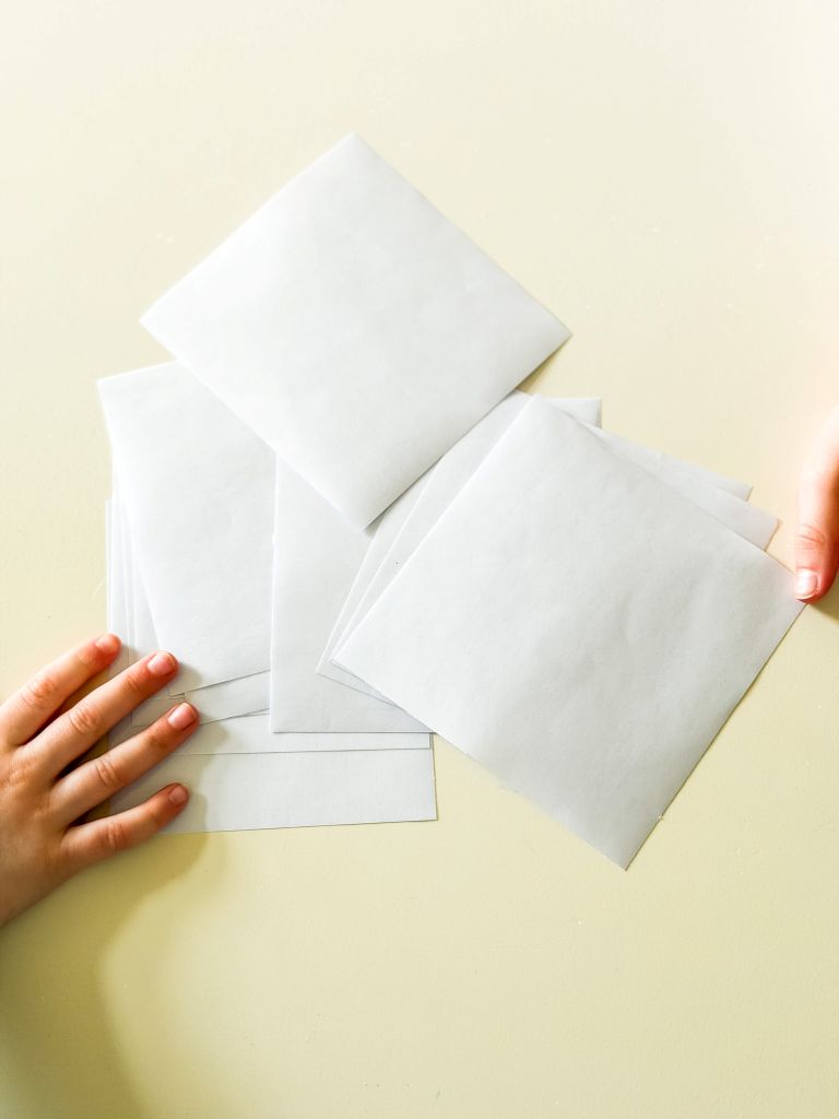 A pile of square-cut white contact paper sheets on a light surface, with a child’s hands resting on either side, ready to begin the base of a DIY pressed flower suncatcher craft.