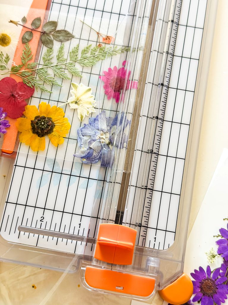 A close-up, top-down view of a clear Fiskars paper trimmer being used to cut transparent sheets layered with colorful pressed flowers, including yellow zinnias, pink cosmos, and green ferns, for a DIY suncatcher project.
